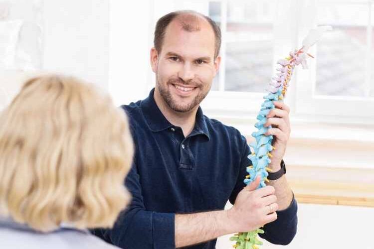 neue-abteilung-fuer-konservative-neurochirurgie-rueckenschmerzen Arzt mit Blick zur Patientin, der eine Modell der Wirbelsäule in der Hand hat